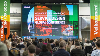 Service Design Global Conferenceの会場風景。参加者の前には大きなスクリーンがあり、イベントのメインビジュアルが映し出されることで、催しが開催される雰囲気を醸成している。