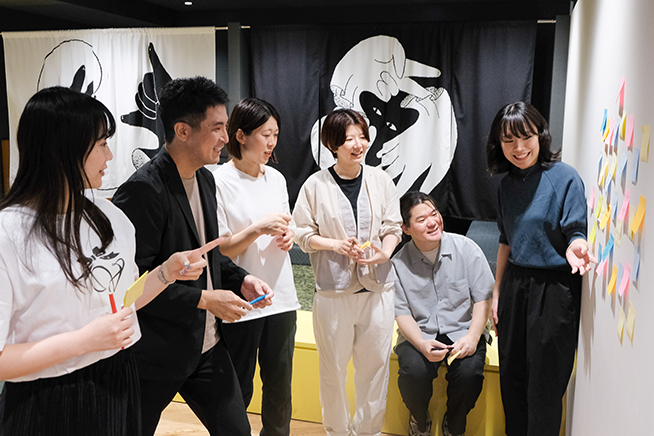 学びの特徴「自分たちの視点をアップデートする」をイメージした写真。様々な世代のメンバーが付箋の貼られたボードの前で意見を交換している様子。