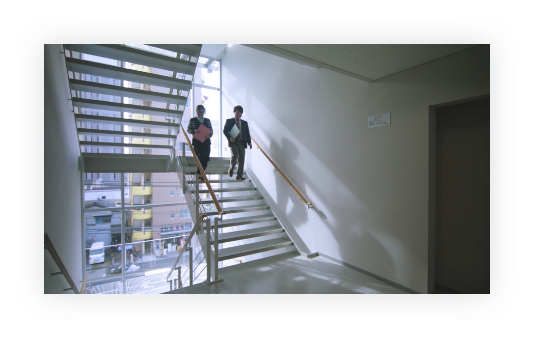 制作した動画のキャプチャ（8枚中2枚目）：富安社内の様子。階段を降りてくる社員2名の姿がある。