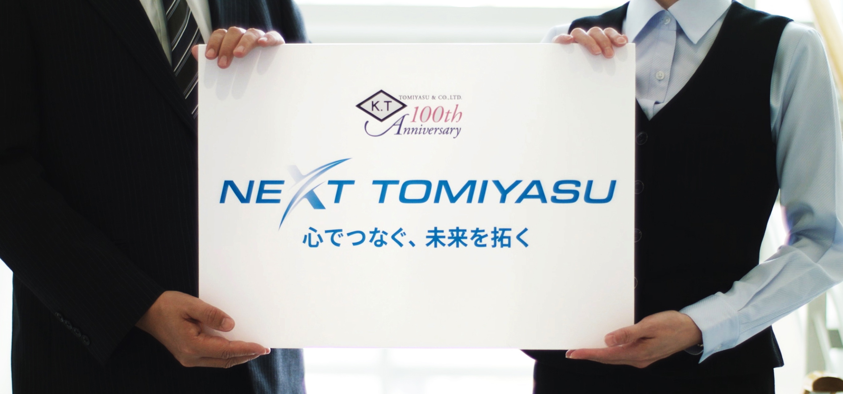 写真：白いパネルを持った富安の社員2名。パネルには100周年のロゴと「NEXT TOMIYASU　心でつなぐ、未来を拓く」というスローガンが書かれている。