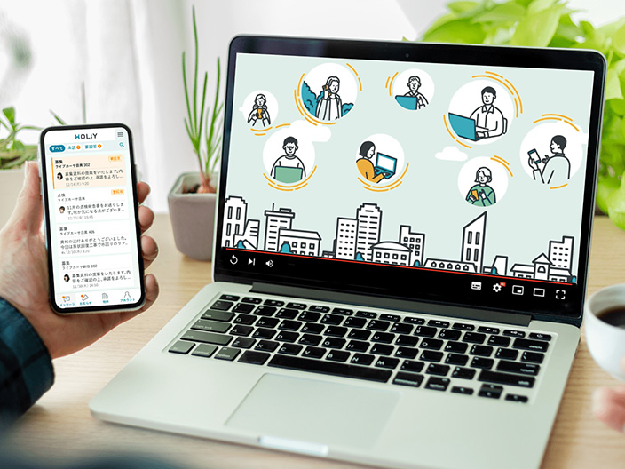 画像：トップのイメージ。スマホとPCの画面のアップが写されている。 スマホにはHOLiYのサービスのUI画面、PCにはサービス紹介動画が表示されている。