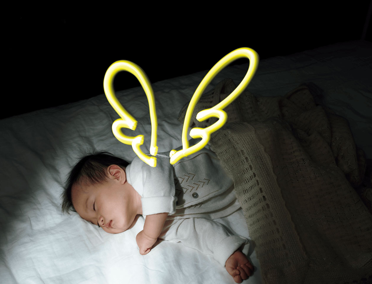 9枚中5枚目：作例写真の一部抜粋。ライブコンポジットというカメラの機能を使って撮影された写真。寝ている赤ちゃんの背中に光の線で羽が描かれている