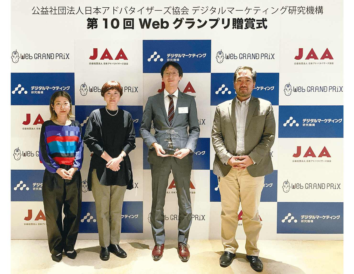 贈賞式での写真。「公益財団法人日本アドバタイザーズ協会 デジタルマーケティング研究機構　第10回Webグランプリ贈賞式」という文字とロゴが入ったバックパネルを背に、贈賞式に参加した亀田製菓様とコンセントのメンバー4名が並んで立っている。