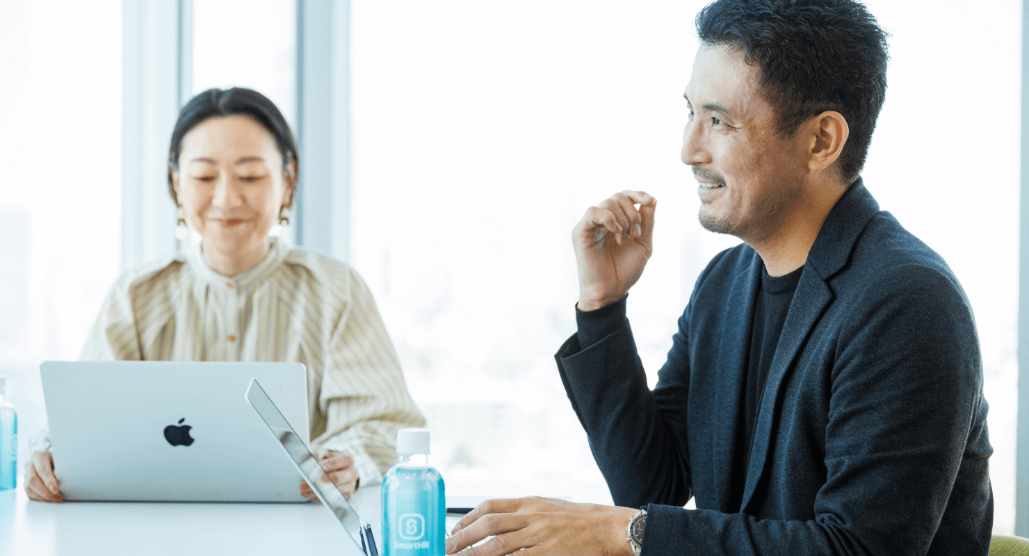 写真:大崎優が話している様子。後ろで本間が笑顔を浮かべている。