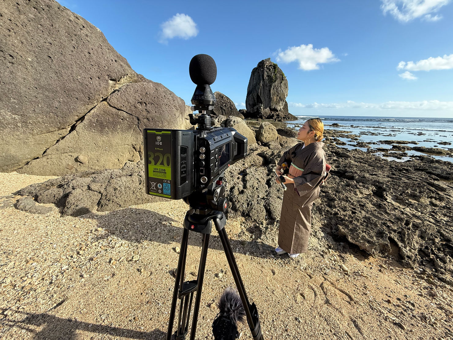 コンセントのイマーシブ映像チーム「渡邊課」が撮影ディレクションを手がけたイマーシブコンテンツ『KUSUDA Riko / YOISURA BUSHI & YACHABO BUSHI』の干潟での撮影風景。Blackmagic URSA Cine Immersive越しに、唄者・楠田莉子さんが奄美の海へ向かい、三味線の弾き語りを行う様子が捉えられている。