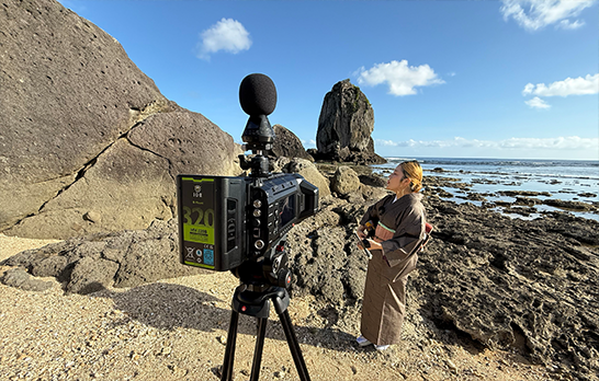 コンセントのイマーシブ映像チーム「渡邊課」が撮影ディレクションを手がけたイマーシブコンテンツ『KUSUDA Riko / YOISURA BUSHI & YACHABO BUSHI』の干潟での撮影風景。Blackmagic URSA Cine Immersive越しに、唄者・楠田莉子さんが奄美の海へ向かい、三味線の弾き語りを行う様子が捉えられている。