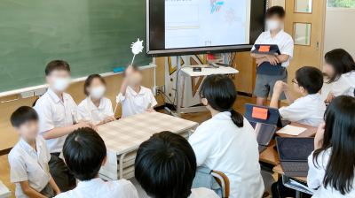 メインビジュアル。教室の黒板の前で児童4名が発表をしている。それを複数の児童が聞いている。