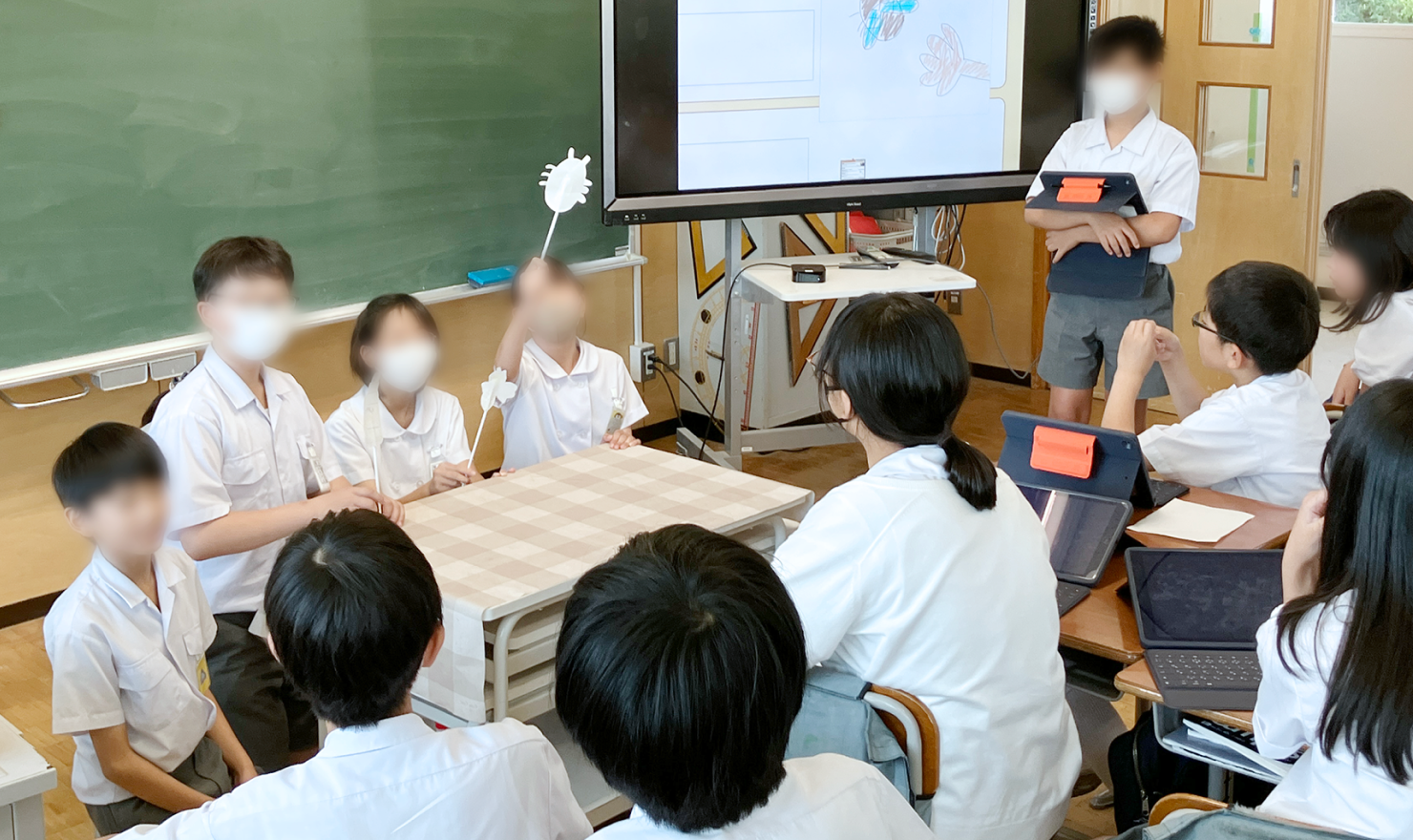 メインビジュアル。教室の黒板の前で児童4名が発表をしている。それを複数の児童が聞いている。