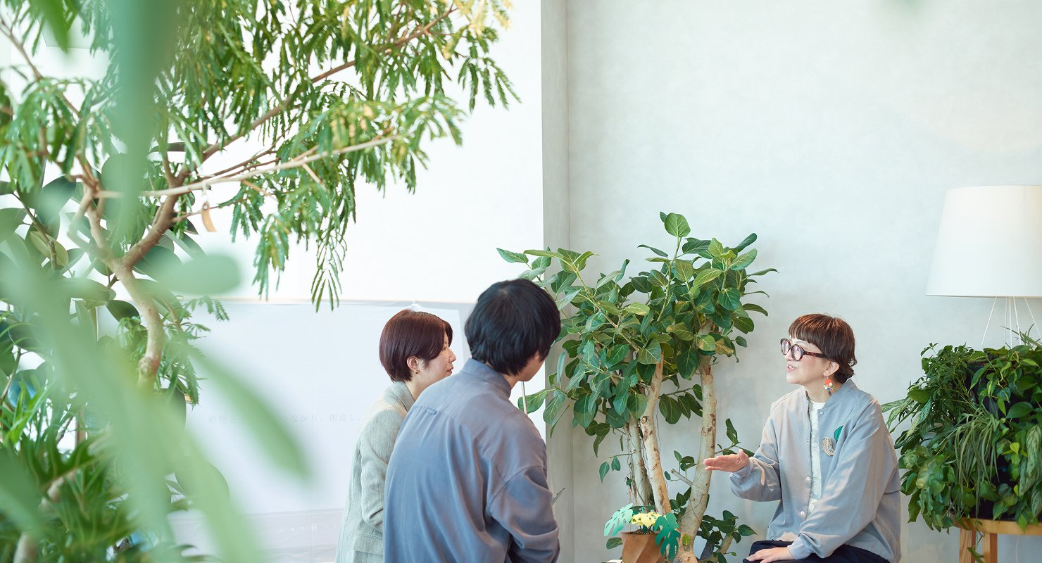 観葉植物に囲まれた店内で、3名が会話をしている様子。